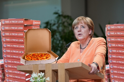 Chancellor Angela Merkel of Germany displays her free pizza – an apology from NSA for monitoring her cell phone.  The NSA also offered to kill one person for her.